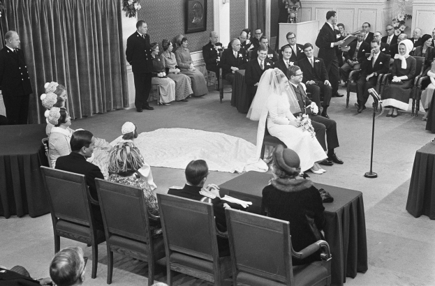 Schwarze und weiße Fotografie einer Hochzeitszeremonie mit sitzenden und stehenden Menschen, ein Mann mit Mikrofon, eine Frau mit einem Strauß, ein Podium mit Mikrofon, Blumenschmuck, ein Vorhang, ein gerahmtes Foto an der Wand und eine Tür im Hintergrund.