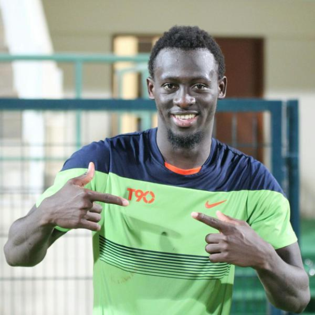 Ein Mann in einem gr├╝nen und blauen Hemd, identifiziert als Emmanuel Adebayor, zeigt auf die Kamera und l├Ąchelt, mit einer unscharfen Gel├Ąnder, Wand und T├╝r im Hintergrund.