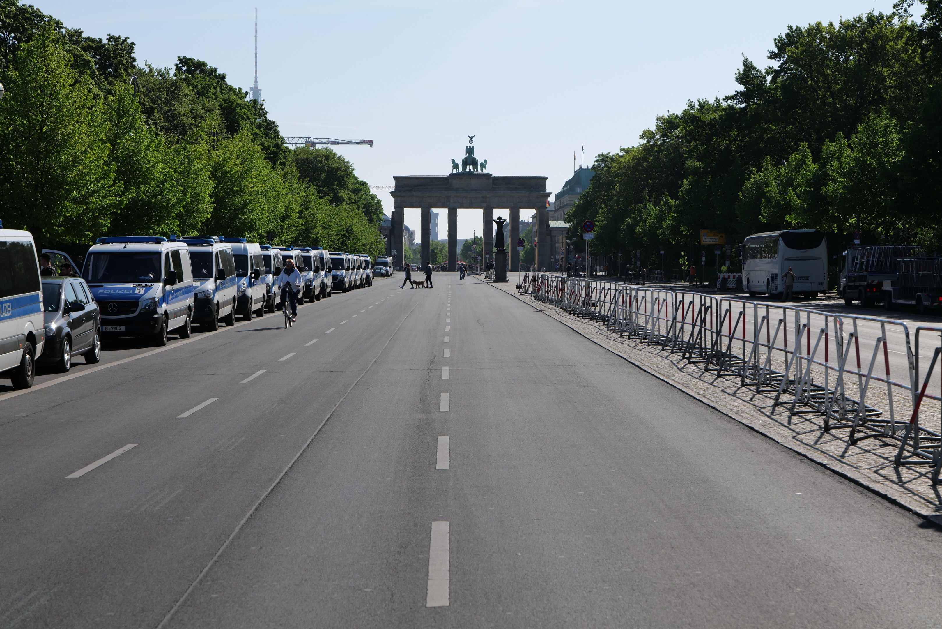 Lange Reihe von Polizeiwagen, die an einer Straße vor dem Brandenburger Tor in Berlin geparkt sind, mit Fahrradfahrern, Fußgängern, Barrieren und Bäumen; ein Bogen mit Statuen ist im Hintergrund zu sehen.