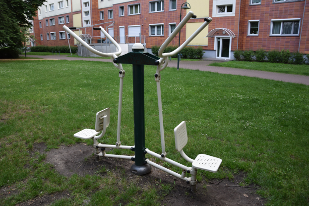 Outdoor-Gym-Geräte in einer grünen Fläche umgeben von Bäumen und Pflanzen, mit Gebäuden im Hintergrund.