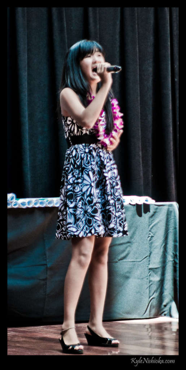 Eine Frau in einem bunten Kleid singt auf der Bühne in ein Mikrofon, mit einem Tisch mit Papieren und einem Vorhang im Hintergrund.