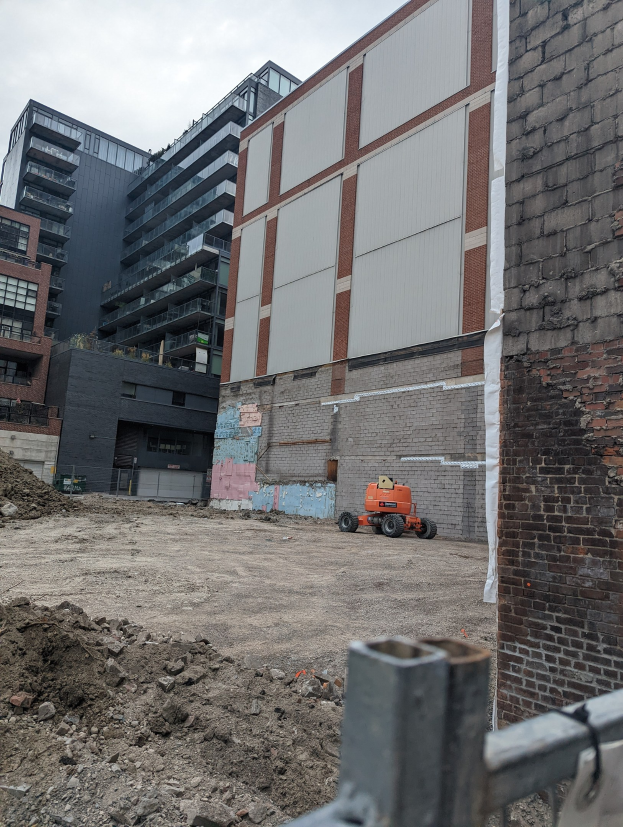 Baustelle mit einem gro├čen, abbruchreifen Geb├Ąude, einem Fahrzeug, einem Metallzaun und einem bew├Âlktem Himmel.