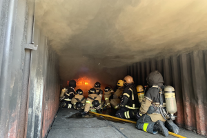 Feuerwehrleute in Schutzausrüstung arbeiten in einem raucherfüllten Tunnel mit einem sichtbaren Feuer im Hintergrund, einer hält einen Schlauch.