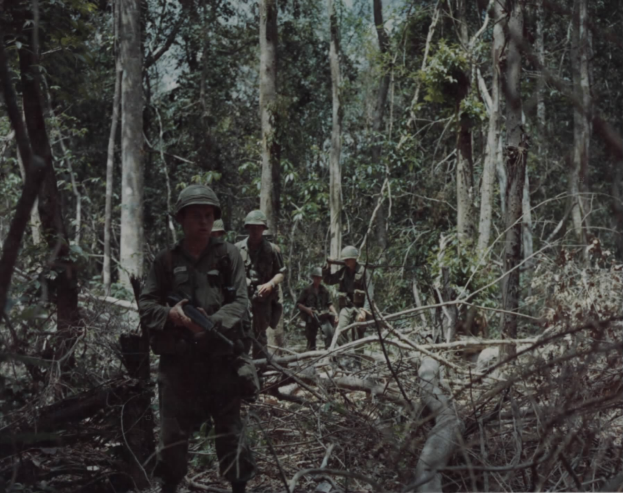 Eine Gruppe von Soldaten in Helmen und mit Gewehren marschiert durch einen dichten Wald mit hohen Bäumen, mit Text am unteren Bildrand.