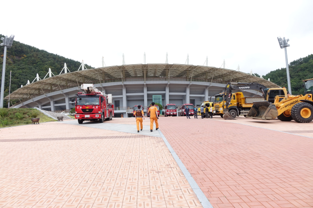 Eine Gruppe von Menschen steht vor dem Olympischen Stadion in Seoul, Südkorea, mit Fahrzeugen auf der Straße, Gras und einer Bank links, Bäumen im Hintergrund und einem klaren blauen Himmel.