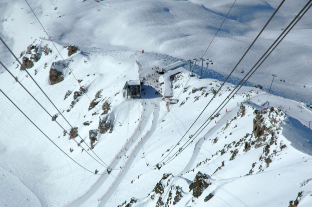 Eine Luftaufnahme eines Skilifts, der einen schneebedeckten Berg hinabfährt, mit Seilbahnkabeln, Felsen und Häusern im Hintergrund, bei strahlendem Sonnenschein.