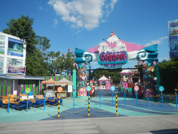 Kirmesattraktion am Parkeingang mit Straße, eingezäuntem Bereich, Buden, Schildern, Bäumen und einem bewölkten Himmel im Hintergrund.