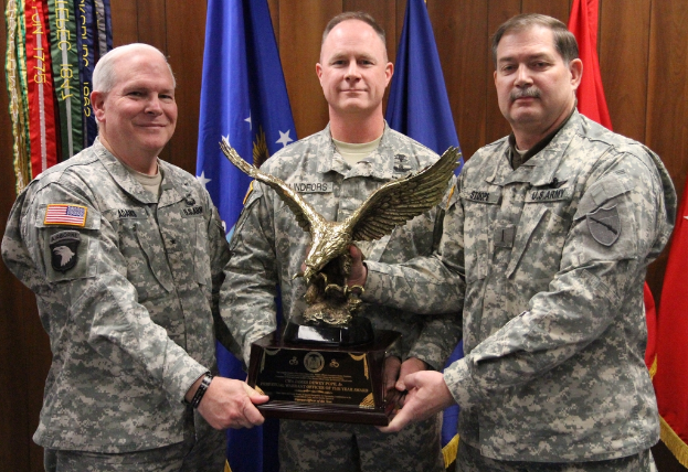 Drei Männer in Uniformen halten Auszeichnungen, mit Fahnen im Hintergrund und bunten Text in der oberen linken Ecke.