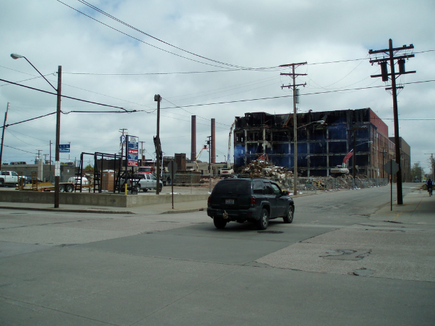Ein Fahrzeug auf der Straße mit einer Laterne mit Kabeln auf der linken Seite und einer ähnlichen Laterne auf dem rechten Gehweg, Gebäude, andere Fahrzeuge und Wolken im Hintergrund.