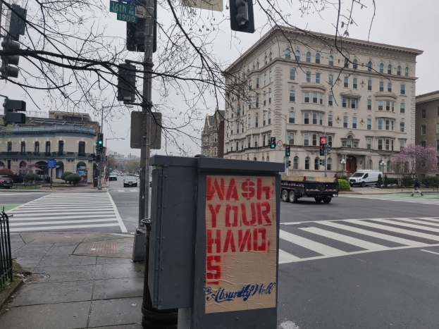 Eine belebte Stadtstraße mit Fahrzeugen, einem Fußgänger, einem Handwaschsymbol auf einem Müllcontainer, Verkehrsampeln, Schildern, einem Metallzaun, Grünpflanzen, Gebäuden und einem klaren Himmel.