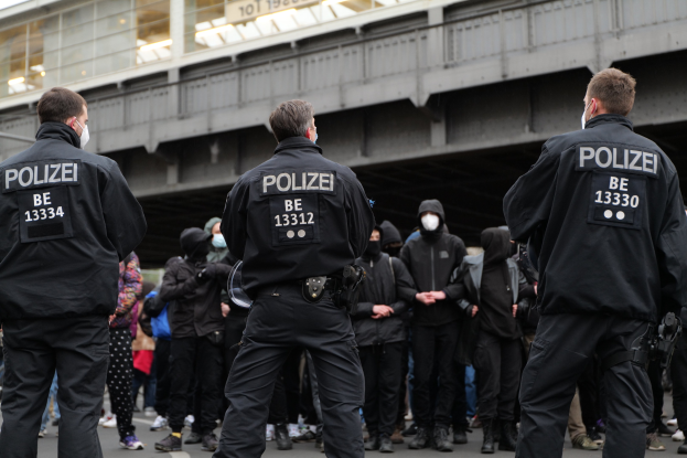 Polizisten in schwarzen Uniformen und Masken vor einer Menge stehend, mit einer Brücke und einem Gebäude im Hintergrund.