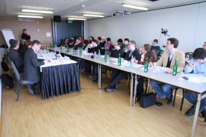 Ein Büroraum mit Angestellten an Tischen sitzend, Flaschen und Papieren auf den Tischen, Holzfußboden, Rucksäcke, ein stehender Mann im Hintergrund, eine Kamera, Lautsprecher, Schilder, Projektor, Deckenlampen, ein schwarzer Vorhang und viele Männer in Anzügen.