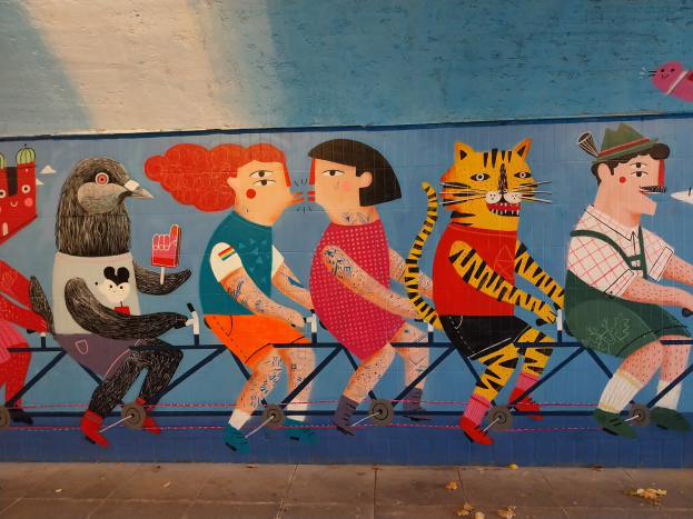 Mural auf einer Gebäudeseite, das Menschen beim Radfahren zeigt, mit einer Straße darunter und Himmel darüber, Teil einer Londoner Street-Art-Serie.