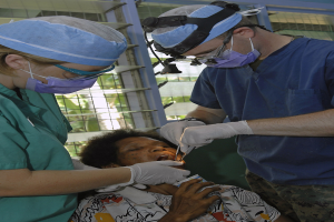 Zwei Chirurgen in Operationskleidung führen eine Gesichtsoperation an einer Frau durch, die auf einem Bett liegt, mit einem Fenster und Bäumen im Hintergrund.