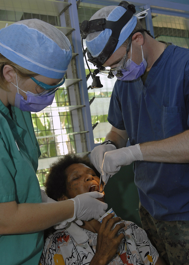 Zwei Chirurgen in Operationskleidung führen eine Gesichtsoperation an einer Frau durch, die auf einem Bett liegt, mit einem Fenster und Bäumen im Hintergrund.