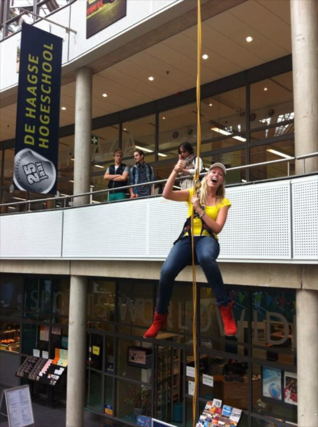 Eine Frau in einem gelben T-Shirt, einer Mütze und roten Stiefeln hängt an einem Seil in einem Geschäft, das wie eine Buchhandlung aussieht, mit Regalen voller Bücher, einem Banner, ein paar Menschen, Glastüren und Deckenleuchten im Hintergrund.