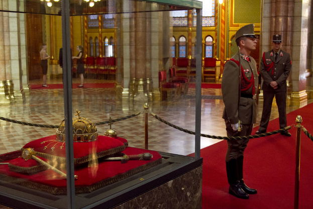 Fünf Personen auf Teppichen mit Stangen, Seilen und einem durchsichtigen Glasbehälter links, einer Säule, einem Licht, Stühlen und einer Wand dahinter sowie Kronleuchtern über drei der Personen.