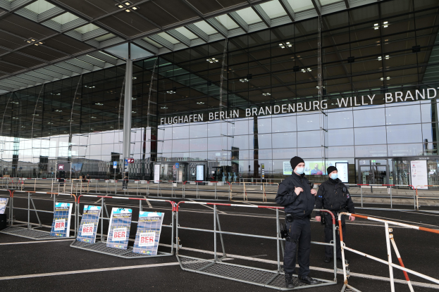 Zwei Polizisten in schwarzen Uniformen stehen vor einem großen Glasgebäude mit Text darauf, mit Absperrungen und Schildern im Vordergrund.