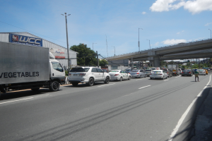 Eine vielbefahrene Straße mit Autos und Lastwagen, eine Brücke im Hintergrund, Laternenpfähle, Bäume, Gebäude und ein bewölkter Himmel.