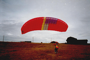Eine Person gleitet mit einem roten Fallschirm über ein Feld, umgeben von Häusern, Pfählen und einem klaren blauen Himmel.