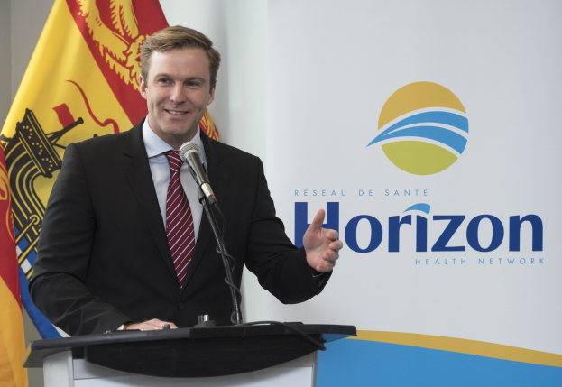 Ein Mann mit einem freundlichen Gesichtsausdruck in einem schwarzen Blazer, einem weißen Hemd und einer roten Krawatte steht an einem Podium mit einem Mikrofon und spricht als Chief Executive Officer.