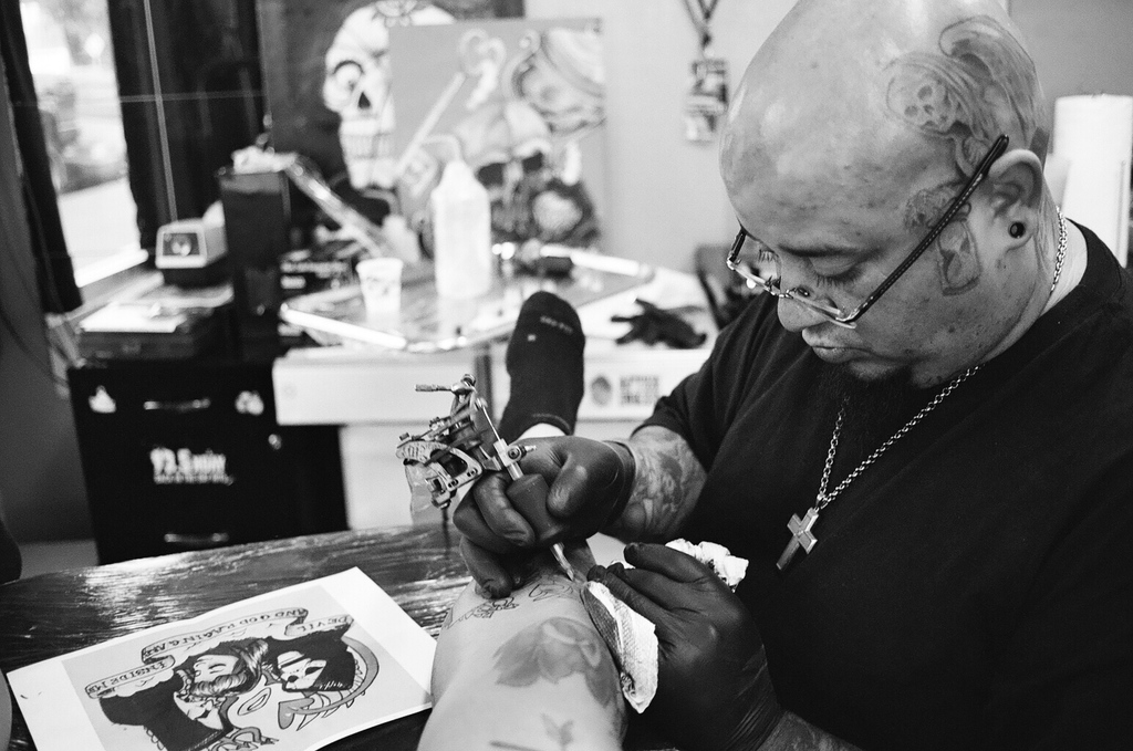Ein Schwarz-Weiß-Bild eines Mannes mit einem schwarzen T-Shirt, einer Brille und einer Kette um den Hals, der mit einer Maschine ein Bein tätowiert, mit einem Blatt auf der Oberfläche und einem unscharfen Hintergrund mit einer Flasche, einem Becher und anderen Gegenständen.
