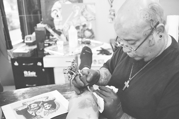 Ein Schwarz-Weiß-Bild eines Mannes mit einem schwarzen T-Shirt, einer Brille und einer Kette um den Hals, der mit einer Maschine ein Bein tätowiert, mit einem Blatt auf der Oberfläche und einem unscharfen Hintergrund mit einer Flasche, einem Becher und anderen Gegenständen.