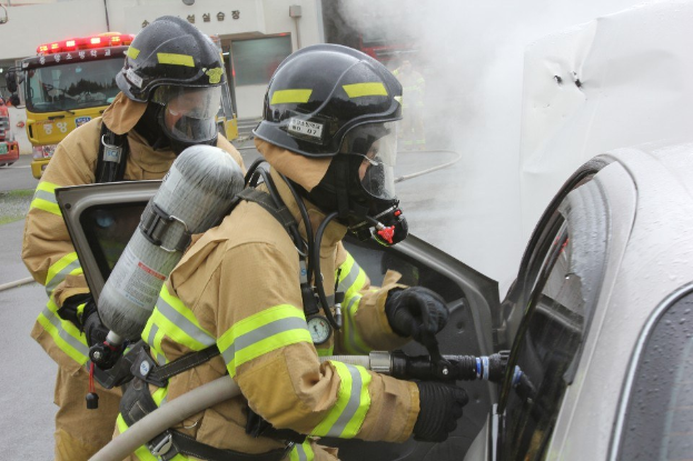 Feuerwehrleute in Schutzausrüstung löschen ein brennendes Auto mit einem Schlauch, während Rauch aus dem Fahrzeug aufsteigt; andere Autos und ein Gebäude sind im Hintergrund zu sehen.