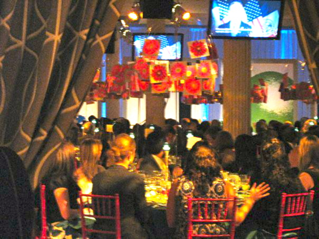 Eine Gruppe von Menschen sitzt an Tischen mit Essen, Fernsehern, Lichtern, hängenden Papieren und Vorhängen in einem Raum, der wie eine Party aussieht.