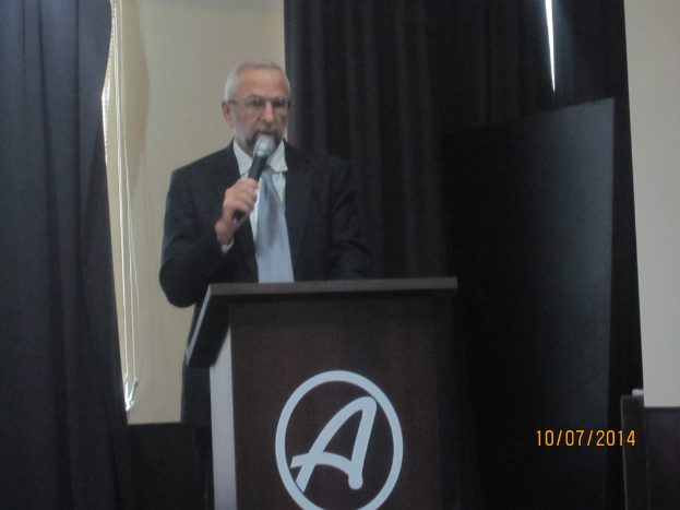 Eine Person in einem schwarzen Mantel und grauer Krawatte steht an einem Schreibtisch und hält ein Mikrofon, während sie eine Rede hält, mit schwarzen Vorhängen im Hintergrund.