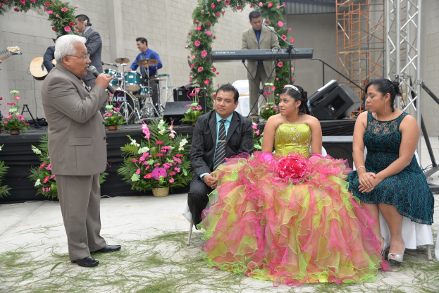 Ein Mann steht an einem Mikrofon und spricht, während ein Orchester im Hintergrund spielt, auf einer Bühne, die mit Blumen und Pflanzen geschmückt ist, mit einigen Menschen im Vordergrund sitzend und anderen stehend.