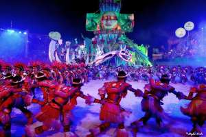 Eine Gruppe von Menschen in farbenfrohen Kostümen tanzt auf einer Bühne vor einem sitzenden Publikum, mit Lichtern, Pfählen, Ballons und einem Bildschirm, der eine Person in der Hintergrund zeigt, während des Karnevals in Rio de Janeiro 2014.