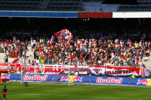 Ein Fußballspiel in einem Stadion mit Spielern auf dem Feld, ein Torpfosten mit Netz, Banner, ein Metallzaun, eine Anzeigetafel, ein Anzeigebildschirm und ein Dach mit Deckenbeleuchtung.