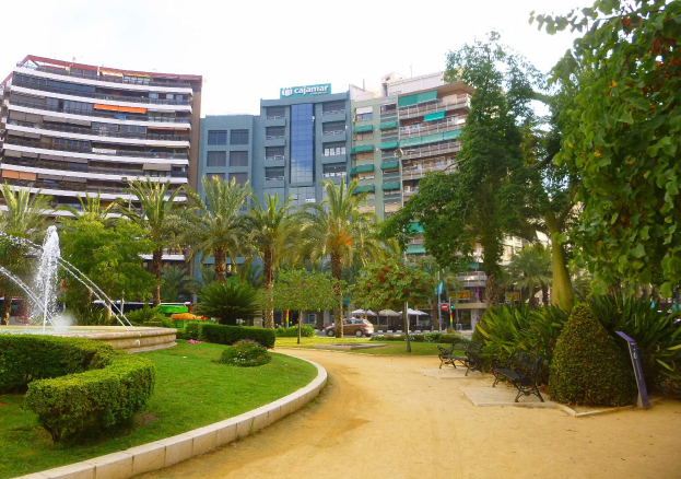 Ein Park mit einem zentralen Brunnen, umgeben von grünem Gras, Pflanzen, Bäumen, Bänken und Laternen, mit Fahrzeugen auf einer nahen Straße und hohen Gebäuden unter einem klaren blauen Himmel im Hintergrund.