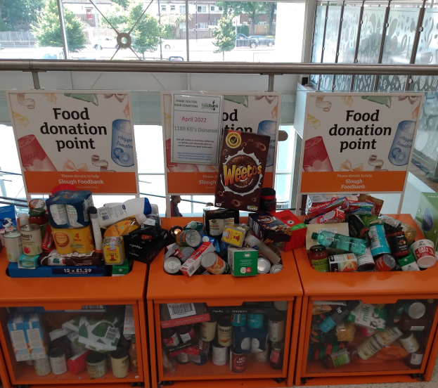 Orange Schübe mit Lebensmittelspenden gefülllt, darunter Flaschen, Dosen und Kisten, mit informativen Schildern im Hintergrund; Glasfenster im Hintergrund zeigen Fahrzeuge, Bäume, Gebäude und Himmel.