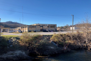 Ein Fluss fließt durch ein grünes Feld neben einem Gebäude neben Bäumen, Pflanzen, Gras, Steinen, Straßenlaternen, Strommasten, Kabeln, Hügeln und einem klaren blauen Himmel, wobei das Gebäude für eine Wasseraufbereitungsanlage geplant ist.