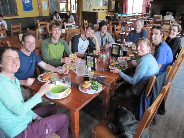 Eine Gruppe von Menschen sitzt um einen Tisch, der mit Tellern, Schüsseln, Gläsern, Flaschen, einem Wasserglas, Karten, Tüchern und Essen gefüllt ist, und lacht und unterhält sich, während ein Jackett über einen der Stühle gehängt ist, und im Hintergrund noch mehr Menschen an anderen Tischen sitzen, mit einem Banner, Bildern und Plakaten an der Wand und einem Fenster mit Vorhängen.