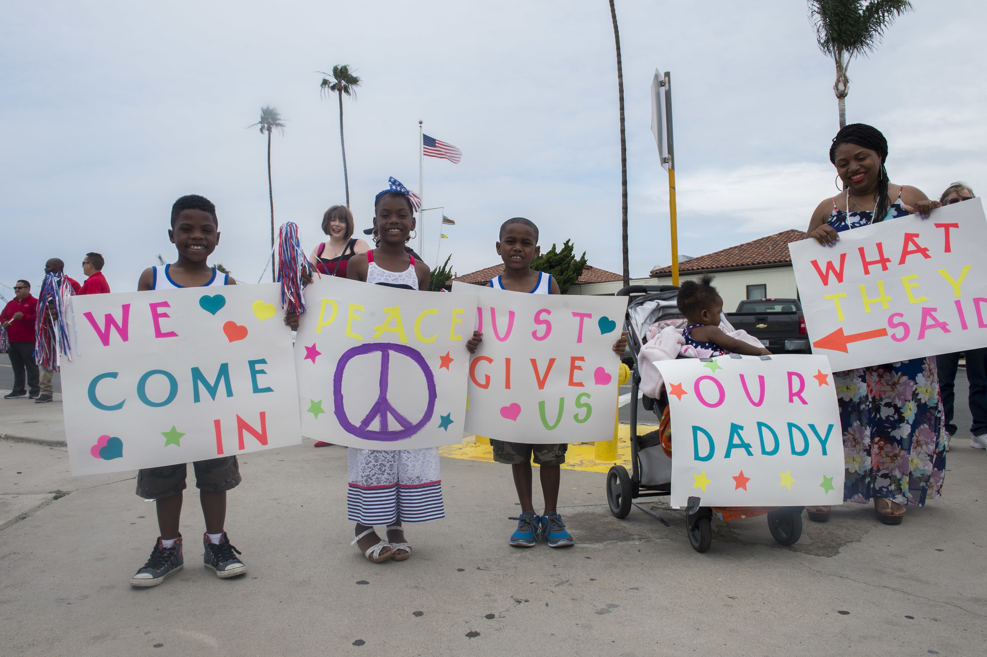 Eine Gruppe von Kindern hält Schilder in der Hand und steht vor einem Gebäude, während eine Frau einen Kinderwagen schiebt, in dem ein Baby liegt; auf einem Schild steht 'Wir kommen in Frieden, gebt uns unseren Daddy', umgeben von Bäumen, Strommasten, einer Flagge, einem Haus, Pflanzen und einem bewölkten Himmel.