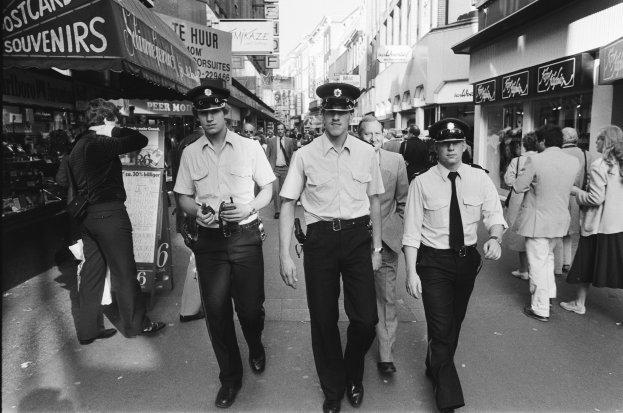 Drei Polizeibeamte in Uniform gehen eine belebte Stadtstraße mit hohen Gebäuden, Geschäften und Passanten entlang.