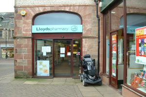 Eine Apotheke mit einem Fahrzeug davor und einem Gebäude links daneben.