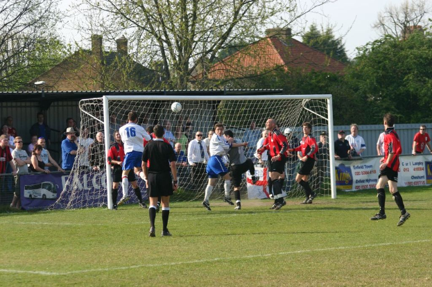 Spieler spielen Fussball auf einem Feld mit einem Tor und Zuschauern dahinter, mit Bäumen und Häusern im Hintergrund.