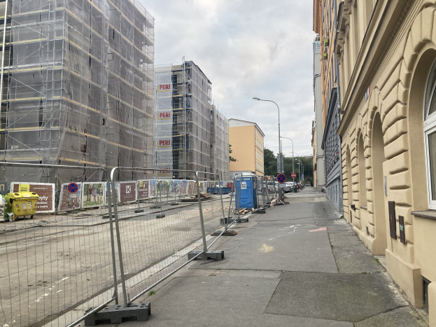 Stadtstra├če mit Geb├Ąuden, Fahrzeugen, Stra├čeninfrastruktur, B├Ąumen und einer Baustelle mit Gerüst im Vordergrund.