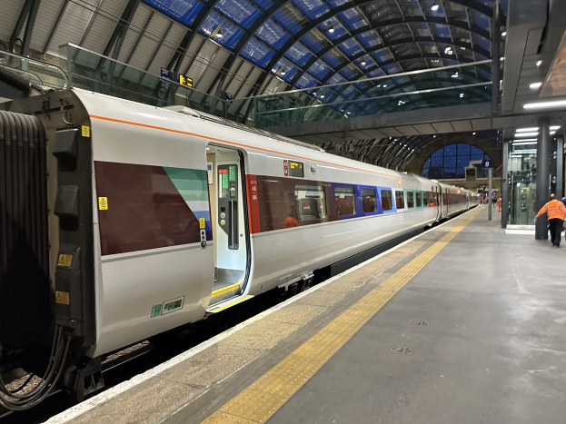 Ein Zug fährt in einen Bahnhof ein, mit Menschen auf dem Bahnsteig und einem Müllcontainer im Vordergrund sowie Deckenlampen im Hintergrund.