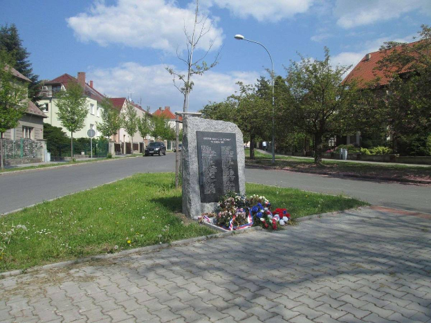 Ein Gedenkstein in einer Straße, umgeben von Gras, Pflanzen, Bäumen, Laternenmasten und Fahrzeugen, mit Gebäuden und einem bewölkten Himmel im Hintergrund, gewidmet den Holocaust-Opfern.