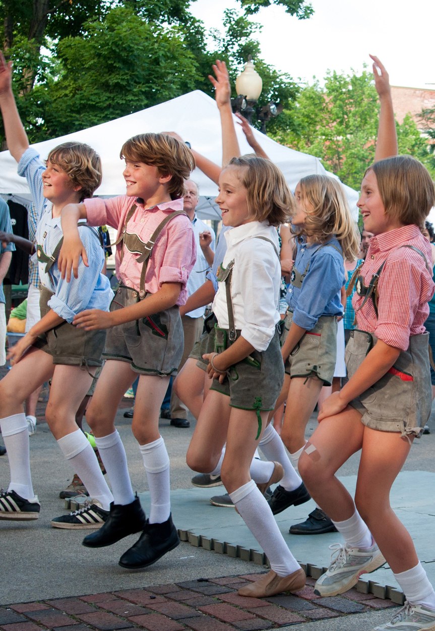 Eine Gruppe von Kindern in traditioneller bayrischer Tracht tanzt auf einer Straße, umgeben von einer Menge, Zelten, Laternenmasten, Bäumen, Gebäuden und einem klaren blauen Himmel, wobei einige Kinder Shorts und Schuhe tragen.