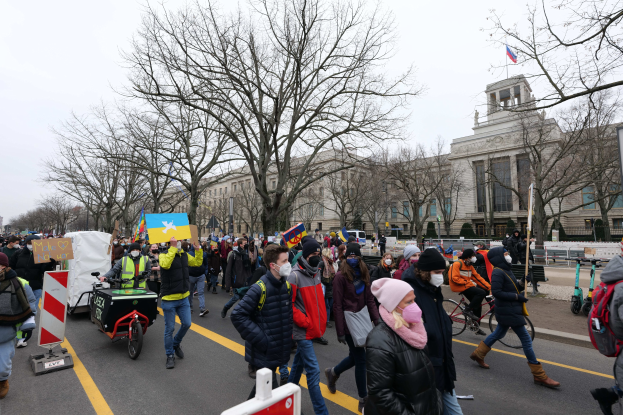 Eine große Gruppe von Menschen marschiert auf einer Demonstration in Washington, D.C. entlang einer Straße, wobei sie Schilder und Banner schwenken und einige Fahrräder fahren, unter einem klaren blauen Himmel.