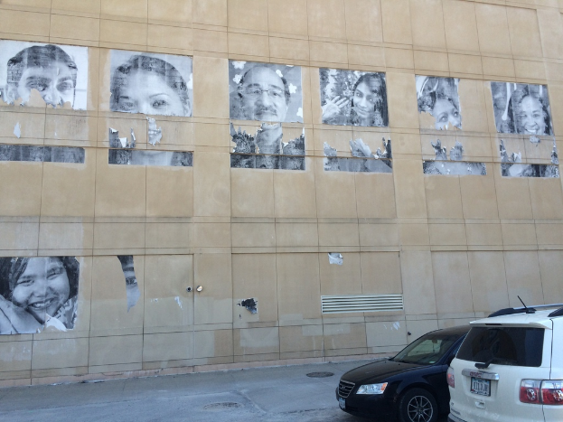 Ein Gebäude mit Autos und Gullis auf der Straße im Vordergrund und Plakate von Menschen an einer Wand im Hintergrund.