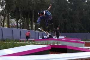 Ein Skateboarder führt einen Trick in der Luft auf einer Rampe aus, umgeben von einer Gruppe von Menschen, die auf dem Boden stehen, einige halten Skateboards, mit Taschen, Flaschen und anderen Gegenständen, die herumliegen, Gras, eine Wand, Bäume, Gebäude und einen klaren blauen Himmel im Hintergrund.