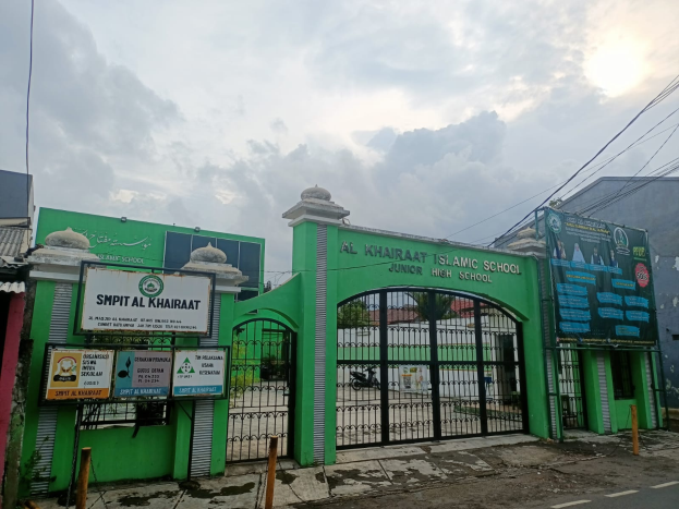 Der Eingang der Al Khairat Islamischen Schule, mit einem gr├╝nen Tor und einer Tafel darauf. Die Stra├če ist unten im Bild zu sehen, und es gibt ein paar Pfosten und Bretter am Tor. Im Hintergrund gibt es Geb├Ąude, B├Ąume und einen klaren blauen Himmel.