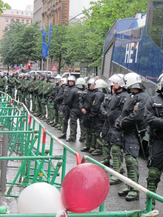 Eine Gruppe von Polizisten in Helmen und mit Waffen in Händen steht vor einer Barriere auf einer Straße mit Geländern und Luftballons, mit Bäumen, Gebäuden, Flaggen und einem klaren blauen Himmel im Hintergrund.